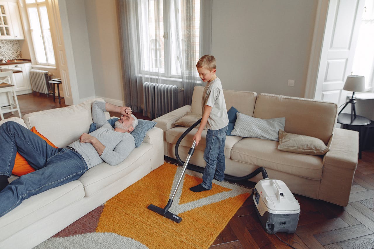 A young boy vacuums the living room carpet while his grandfather relaxes on the sofa.